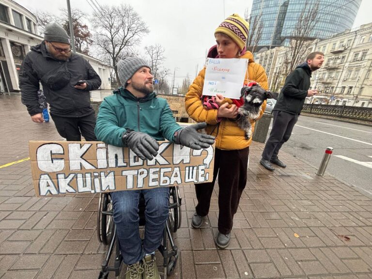 На фото чоловік, який користується кріслом колісним та жінка, яка тримає песика. Вони стоять на тротуарі біля проїзної частини. Чоловік тримає табличку з написом "скільки акцій треба ще?". Жінка тримає папір з написом "ваші 10 секунд = мої 10 хвилин".