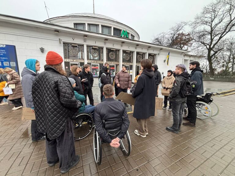 На фото велика група людей, які зібралися в коло. Серед них є люди, які користуються кріслами колісними. За ними вхід до будівлі із позначкою М — метро.